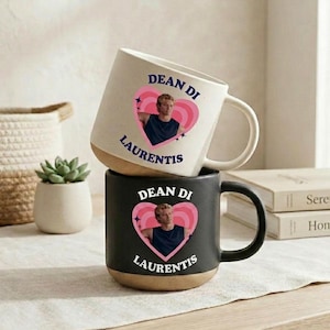 May include: A cream-colored ceramic mug and a black ceramic mug, each featuring a pink heart graphic with a photograph of a person inside, surrounded by the text DEAN DI LAURENTIS in dark blue or white lettering, with a natural tan base.