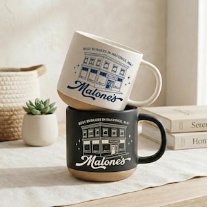 May include: A cream-coloured mug and a black mug, both featuring a blue or white line-art illustration of a building with the text "Best Burgers in Hastings, MA!" and "Malone's" printed on the front. Each mug has a natural, unglazed tan base.