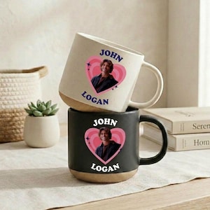 May include: A cream-colored ceramic mug stacked on a black ceramic mug. Both mugs feature a pink heart graphic containing a photograph of a person, with the text JOHN LOGAN printed in blue on the cream mug and white on the black mug.