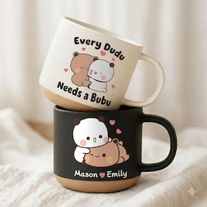 May include: A cream-coloured mug with a speckled finish featuring a brown bear and white bear hugging with the text Every Dudu Needs a Bubu, stacked on a matte black mug featuring a white bear and brown bear with the text Mason and Emily.