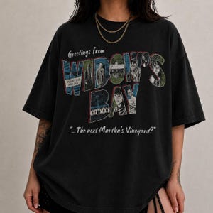 Puede incluir: Una camiseta negra de manga corta con un estampado gráfico que incluye el texto Greetings from WIDOW'S BAY y ...The next Martha's Vineyard? El diseño integra varias imágenes fotográficas pequeñas de personas y edificios dentro de las letras grandes.
