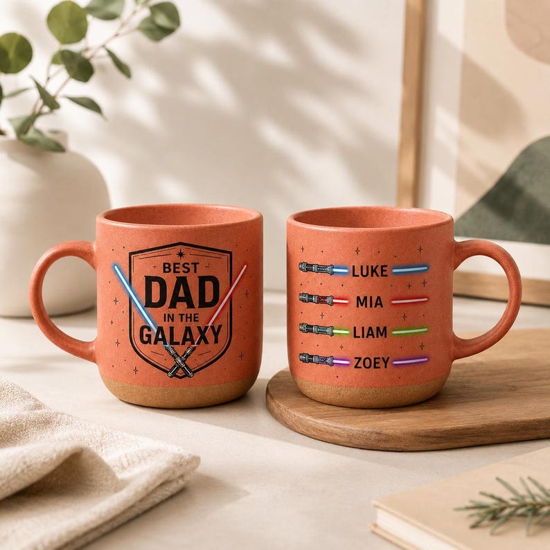 May include: Two orange ceramic mugs with brown bases. One mug has the text "BEST DAD IN THE GALAXY" with lightsaber graphics. The other mug has lightsaber graphics and names: Luke, Mia, Liam, and Zoey. The mugs are on a wooden surface.