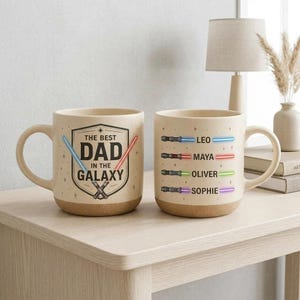 May include: Two beige ceramic mugs with brown bases. One mug has the text "THE BEST DAD IN THE GALAXY" with crossed lightsabers. The other mug has names and lightsaber graphics: Leo, Maya, Oliver, and Sophie.