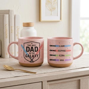 May include: Two pink ceramic mugs with brown bases. One mug has "THE BEST DAD IN THE GALAXY" text and lightsaber graphics. The other mug has names and lightsaber graphics. A gold spoon rests on the table.
