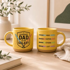 May include: Two yellow ceramic mugs with brown bases. One mug has the text "LEGENDARY DAD IN THE GALAXY" with lightsaber graphics. The other mug has names and lightsaber graphics. The mugs are on a wooden surface.