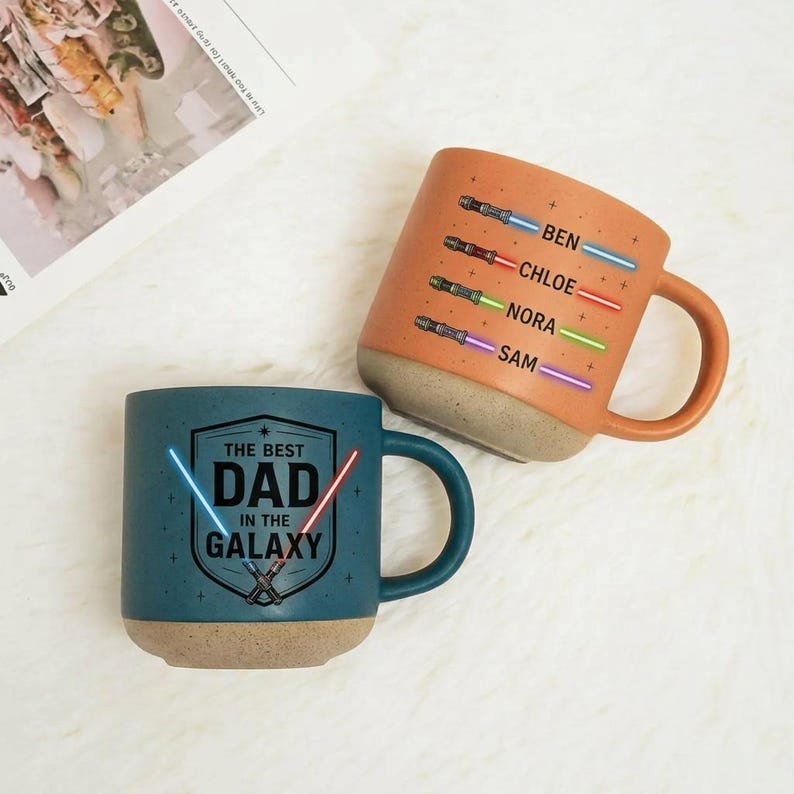 May include: Two ceramic mugs with a space theme. One is teal with "THE BEST DAD IN THE GALAXY" text and lightsaber graphics. The other is orange, featuring names and lightsaber illustrations. Both mugs have a speckled base and a handle.