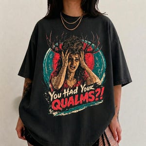 May include: A charcoal gray short-sleeve t-shirt featuring a graphic print of a person with antlers holding their head in distress. The design includes a red circle, teal accents, and the text "You Had Your QUALMS?!" in bold red and white lettering.