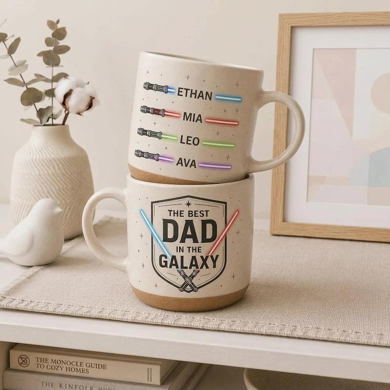 May include: Two beige ceramic mugs stacked on top of each other. The top mug has the names Ethan, Mia, Leo, and Ava with colored lightsaber designs. The bottom mug reads "THE BEST DAD IN THE GALAXY" with lightsaber graphics.