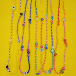 May include: A selection of beaded necklaces in vibrant colours, featuring charms like flowers, hearts, and smiley faces. The necklaces use beads in pink, yellow, blue, and orange, set against a bright yellow backdrop.