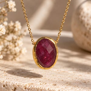 May include: A necklace featuring a faceted, oval-shaped deep red gemstone set in a textured gold-toned bezel. The pendant is suspended from a delicate gold-toned chain, showcasing a polished finish on the stone and a hammered appearance on the metal setting.