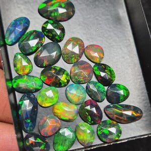 Puede incluir: Una colección de ópalos negros facetados en varias formas ovaladas y de pera. Cada piedra preciosa muestra un color base oscuro con destellos vibrantes de verde, rojo, naranja y azul sobre sus superficies pulidas y multifacetadas.