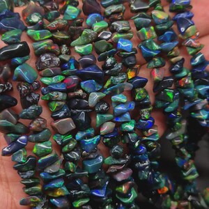 May include: A collection of irregular, tumbled black opal gemstone chips strung together. Each small, polished stone displays a dark base color with vibrant, iridescent flashes of blue, green, and red light reflecting from within the mineral structure.