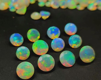 May include: A collection of round, faceted opal gemstones displaying a translucent white base with vibrant flashes of green, blue, orange, and yellow iridescence. The stones are arranged in a cluster on a dark surface, showcasing their play-of-color.