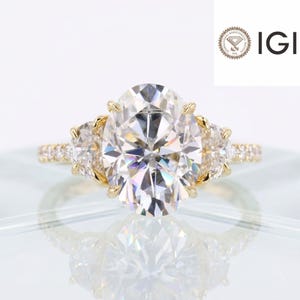 May include: A yellow gold ring featuring a large oval-cut center stone flanked by two smaller half-moon-cut side stones, with a pavé band. The ring is accompanied by an IGI International Gemological Institute certification logo.