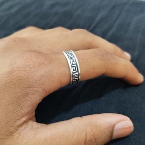 Può includere: Un anello d'argento con un motivo a chiave greca. L'anello, indossato su un dito, presenta un motivo geometrico ripetuto attorno alla sua circonferenza. Il metallo appare lucido.