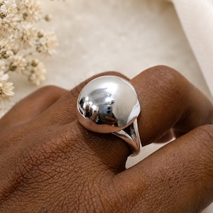 May include: A polished silver ring featuring a large, smooth, dome-shaped centrepiece. The ring has a split-shank band design that connects to the base of the rounded, reflective metallic sphere, creating a bold and clean aesthetic for the finger.