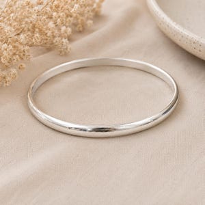 Op de afbeelding: Een zilverkleurige armband met een glad, afgerond ontwerp. De armband wordt gepresenteerd op een neutraal gekleurde stof, met gedroogde bloemen en een keramische plaat op de achtergrond. Een eenvoudig, elegant sieraad.
