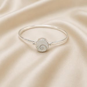May include: A silver bangle bracelet featuring a circular centre stone with a white and grey spiral pattern. The metal band is thin and polished, connecting to the round setting on both sides with small silver spheres, creating a clean design.