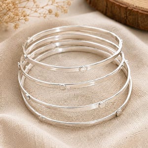 May include: A stack of five silver bangles with small, circular embellishments. The bracelets are displayed on a beige linen surface, with a blurred background featuring dried flowers and a wooden slice.