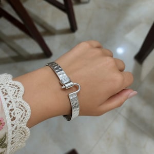 May include: A silver bracelet with a hammered texture and a hook clasp. The bracelet is worn on a person's wrist, showcasing its design and the way it fits. The bracelet's design is simple yet elegant, suitable for everyday wear.