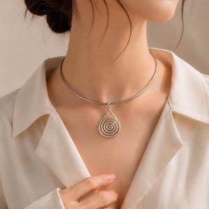 Pode incluir: Um colar prateado com um pendente em forma de gota com um design em espiral. O pendente pende de um fino colar circular. O colar é usado sobre uma camisa creme.
