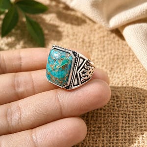 Puede incluir: Anillo plateado con una piedra turquesa cuadrada. El anillo tiene un diseño ornamentado en la banda y un bisel texturizado alrededor de la piedra. La piedra turquesa presenta una mezcla de colores azules y verdes.