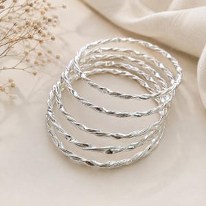 May include: A set of five silver-colored bangles, each featuring a twisted, rope-like texture. The bracelets are circular and have a polished, reflective metallic finish, stacked together to show their uniform design and shiny surface.