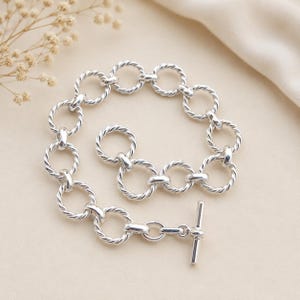 Op de afbeelding: Een zilveren kettingarmband bestaande uit meerdere onderling verbonden cirkelvormige schakels met een gedraaide touwstructuur. De armband is voorzien van een knevelsluiting bestaande uit een gladde zilveren staaf door een grotere ring.