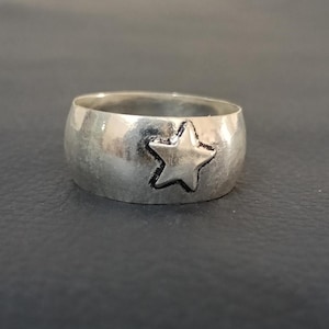 May include: A wide silver-toned band ring featuring a single embossed five-pointed star design on the front. The metal surface has a polished, reflective finish with darkened detailing around the edges of the star shape to provide contrast.