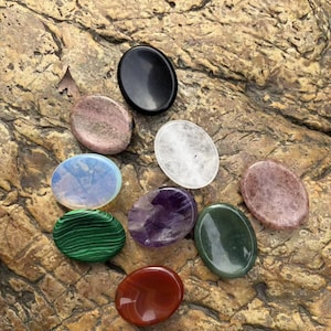 Könnte beinhalten: Eine Sammlung von neun glatten, oval geformten, polierten Steinen in verschiedenen Farben, darunter Schwarz, Weiß, Violett, Grün, Rot und gesprenkelte Rosatöne. Jeder Stein hat eine glänzende Oberfläche und eine abgerundete, flache Form.