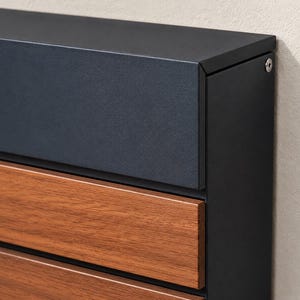 Puede incluir: Un buz&oacute;n de pared con marco de metal negro mate y tapa superior, con paneles horizontales de acabado en tono madera c&aacute;lida. La estructura incluye una cabeza de tornillo circular de color plateado visible en la esquina superior derecha.