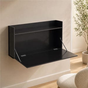May include: A black wall-mounted desk featuring a rectangular frame with an open top shelf and a hinged drop-down panel. The desk is supported by two thin metal support arms attached to the sides, allowing the front panel to fold down horizontally.