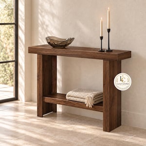 May include: A rectangular dark brown wooden console table with a lower shelf. On the top surface sits a translucent dark glass bowl and two black metal candle holders with lit white taper candles. A folded beige textured throw blanket rests on the shelf.