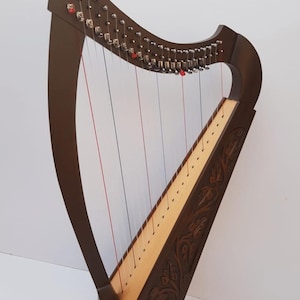 May include: A dark brown wooden harp featuring a light-colored soundboard and intricate floral carvings along the side pillar. The instrument includes silver tuning pins, clear strings, and two distinct red strings positioned at specific intervals.