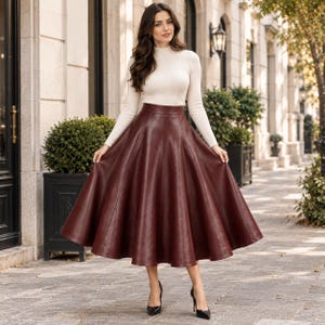May include: A long, high-waisted, burgundy faux leather skirt with a flared, A-line silhouette and a smooth finish, paired with a long-sleeved, cream-coloured mock neck jumper and black pointed-toe high-heeled shoes.