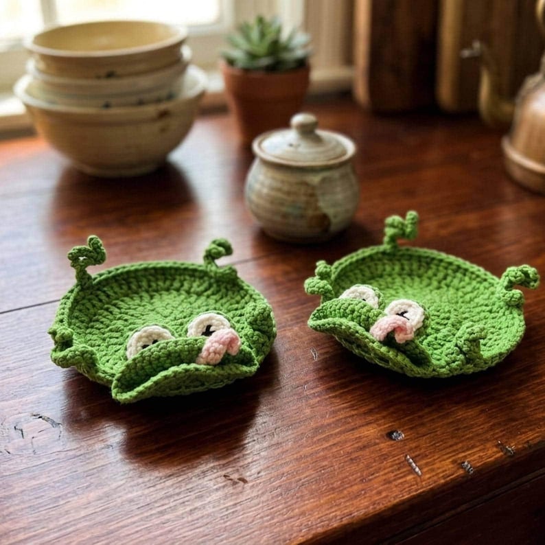 May include: Two round, bright green crocheted coasters shaped like frogs. Each coaster features a textured surface, two white eyes with dark pupils, and a small pink tongue protruding from the centre. The edges are scalloped with small loops.