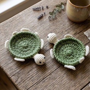 May include: Two handmade crochet coasters shaped like turtles. Each coaster features a circular, textured olive green shell with a raised border and a small, cream-coloured head with two black eyes, along with four cream-coloured flipper accents.