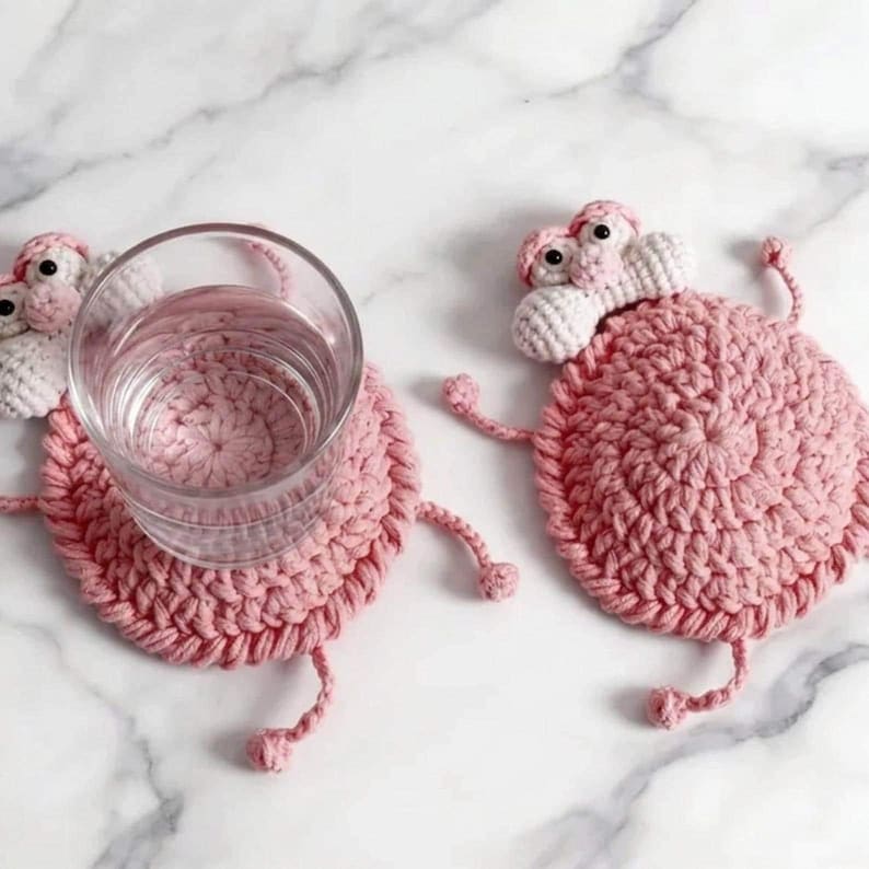 May include: Two round, light pink crocheted coasters shaped like cartoon characters with small white faces, black eyes, and thin yarn limbs. One coaster supports a clear glass tumbler, while the other lies flat, showcasing the textured yarn construction.