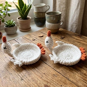 May include: Two handmade crochet swan-shaped bowls crafted from white yarn with orange beaks and feet. Each bowl features a circular base, a raised neck, and small wings, designed to hold small items on a flat surface.