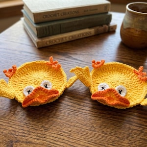 May include: Two round, yellow crochet coasters shaped like ducks. Each coaster features a bright orange beak, small orange feet, and white eyes with black cross-stitched pupils. The circular bodies are crafted with a textured yarn stitch pattern.