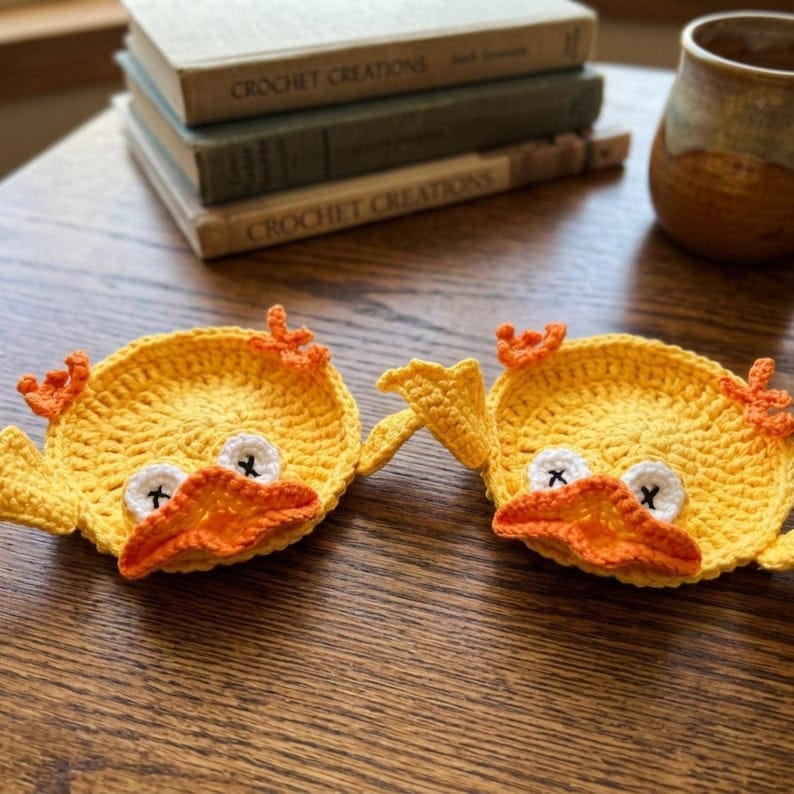 May include: Two round, yellow crochet coasters shaped like ducks. Each coaster features a bright orange beak, small orange feet, and white eyes with black cross-stitched pupils. The circular bodies are crafted with a textured yarn stitch pattern.