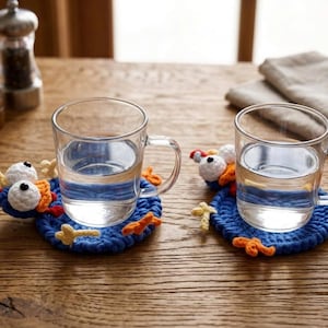 May include: Two clear glass mugs filled with water, each resting on a round, blue crocheted coaster. The coasters feature attached white and orange yarn details resembling cartoon eyes, beaks, and feet, creating a playful, character-like appearance.