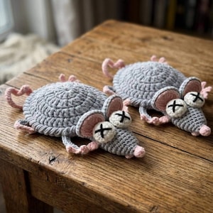 May include: Two handmade crochet mouse coasters featuring a circular grey body, pink ears, a small pink nose, and white eyes with black stitched X marks. Each mouse has a small curled tail and four tiny pink feet extending from the sides.