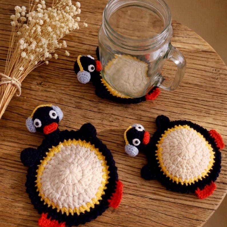 May include: Three handmade crochet coasters shaped like penguins, each featuring a black body, a white circular centre, yellow accents, red feet, and light blue earmuffs. One coaster is positioned under a clear glass mason jar.