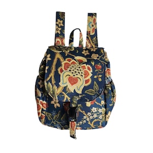 May include: A navy blue backpack featuring a repeating floral pattern in shades of red, yellow, and cream. The bag includes a top flap with a black plastic buckle closure, two shoulder straps, and a small top handle, all made from the same fabric.