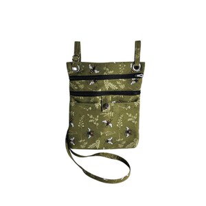 May include: Olive green crossbody bag with a bee and floral pattern. The bag has three zippered compartments and a button closure. It features a long, adjustable strap and metal hardware.