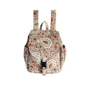 May include: A small backpack featuring an off-white base with a dense floral and paisley pattern in shades of red, orange, and muted green. The bag includes two shoulder straps, a top handle, side pockets, and a front flap secured by a black buckle.