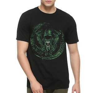 Peut inclure: Un t-shirt noir à manches courtes avec un imprimé graphique circulaire dans des tons verts. Le motif représente une créature détaillée et menaçante avec des dents acérées et une tête allongée, entourée d'un anneau texturé abstrait.