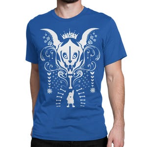 Op de afbeelding: Een koningsblauw t-shirt met korte mouwen en een grote witte grafische print. Het ontwerp toont een gestileerd schedelachtig wezen met een kroon, omringd door ingewikkelde bloemmotieven, sneeuwvlokken en een klein figuurtje onderaan.