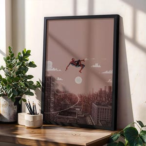 May include: A framed vertical art print featuring a red and blue costumed figure leaping through the air above a detailed cityscape. The illustration uses a muted mauve and brown color palette, depicting buildings, a water tower, and a full moon.